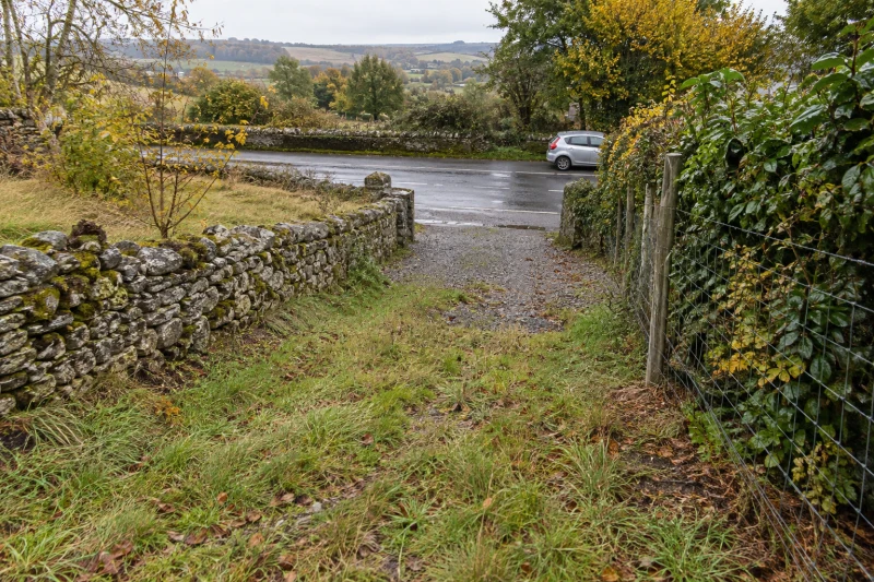 A sloping grassy plot leads down to a narrow gravel entrance and a road.