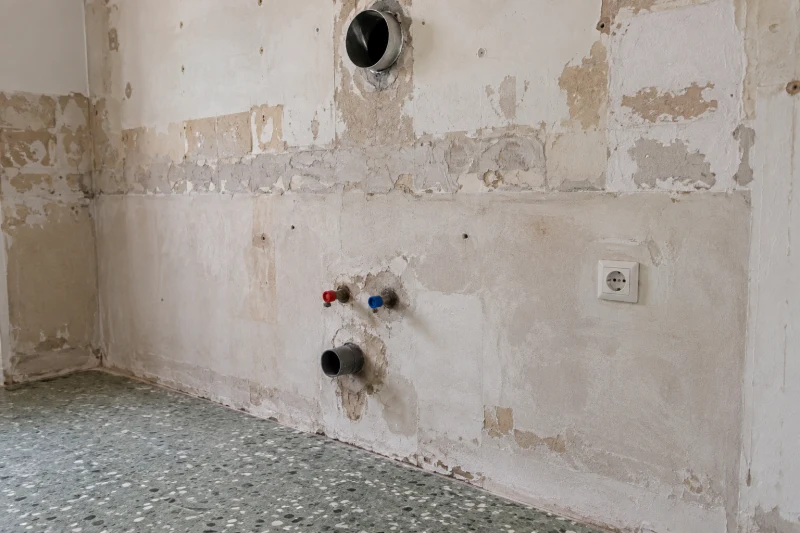 Bare wall in a separate kitchen under renovation, with existing water pipes, drain, electrical outlet and ventilation opening.